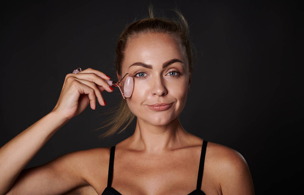 Крупный план европейской женщины средних лет со здоровой светящейся загорелой кожей, массирующее лицо с нефритовым роликом изолированы на черном фоне с копией рекламного пространства - Фото, изображение