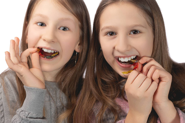 Two cute girls playfully pose with orthodontic appliances in their hands on a white background. The concept of oral hygiene in childhood. Studio photo. - Photo, Image