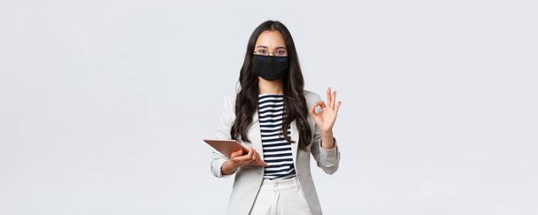 Business, finance and employment, covid-19 preventing virus and social distancing concept. Asian businesswoman with digital tablet, wear protective mask against virus and showing okay sign - Foto, Bild