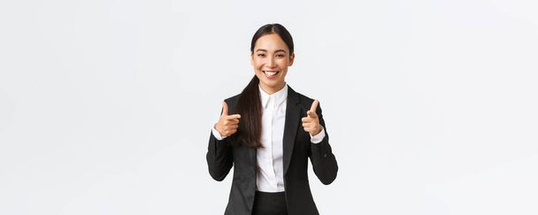 Magabiztos mosolygós ázsiai eladó fekete öltönyben mutatja hüvelykujját, garantálja a termék minőségét vagy a legjobb szolgáltatást. Értékesítési menedzser próbál eladni valamit az ügyfél, fehér háttér - Fotó, kép