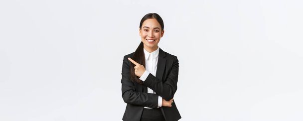 Χαρούμενη επαγγελματίας ασιατική γυναίκα διευθυντής, επιχειρηματίας με κοστούμι δείχνει ανακοίνωση, χαμογελώντας και δείχνοντας το δάχτυλο αριστερά στο προϊόν ή το έργο πανό, στέκεται λευκό φόντο - Φωτογραφία, εικόνα
