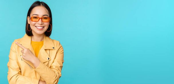 Stylisch lächelnde asiatische Frau mit Sonnenbrille, Zeigefinger nach links, zeigt Werbebanner, steht im trendigen gelben Mantel vor blauem Hintergrund - Foto, Bild
