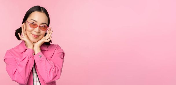 Portrait von stilvollen niedlichen asiatischen Mädchen, lächelnd und berührend Gesicht, Blick nach oben verträumt, nachdenklich Blick, steht über rosa Hintergrund - Foto, Bild