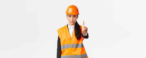 Seriös aussehende selbstbewusste und unzufriedene asiatische Ingenieurin schüttelt den Finger, schimpft Bauarbeiter wegen gefährlichen Verhaltens, verbietet und verbietet Handlungen an Baustellen, weißer Hintergrund - Foto, Bild