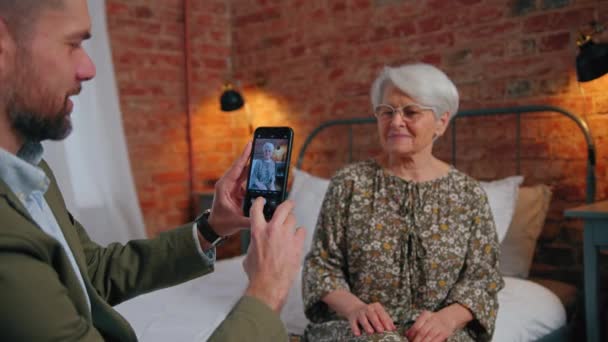 middle-age caucasian entrepreneur taking photo of his elderly grey-haired mother with his smartphone  - Footage, Video