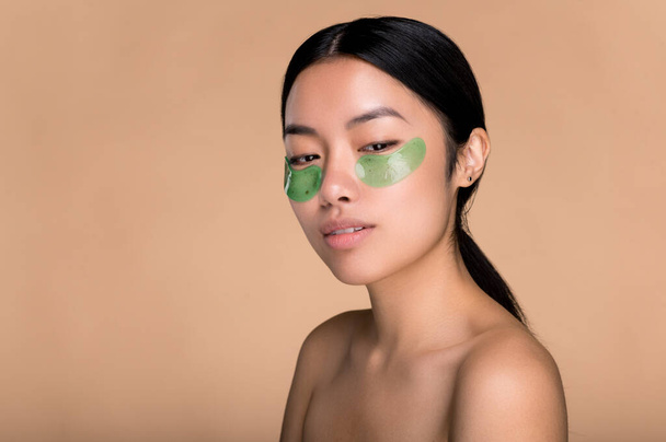 Κινεζική όμορφη κοπέλα με τέλειο δέρμα και γυμνούς ώμους, με καλλυντικά κολλαγόνο πράσινο κηλίδες κάτω από τα μάτια, φροντίζει το δέρμα γύρω από τα μάτια, αποτρέπει τις ρυτίδες, κοιτάζει κάτω, θέτοντας σε απομονωμένο μπεζ φόντο - Φωτογραφία, εικόνα