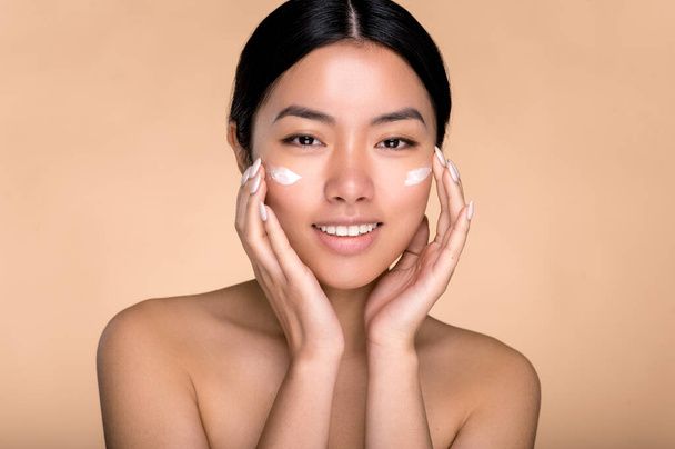Facial cream. Asian attractive girl with perfect facial skin applied moisturizer, poses for the camera with hands near her face, stands on an isolated beige background, looks at the camera, smiles - Photo, Image