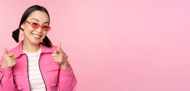 Close up portret nowoczesnej azjatyckiej dziewczyny w okularach przeciwsłonecznych uśmiecha, wskazując palcami na aparat, chwalić, zapraszając lub komplement, stojąc nad różowym tle - Zdjęcie, obraz