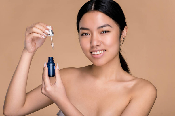 Attractive beautiful asian girl with naked shoulders, holding open face serum in hand, demonstrating moisturizer serum, looking at camera, smiling, stands on isolated beige background - Foto, Imagem