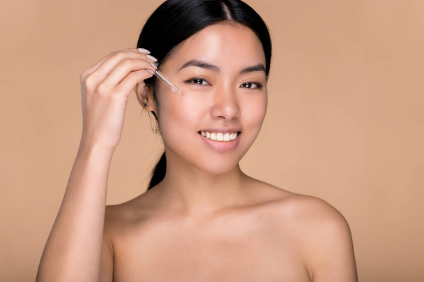 Lovely smiling asian brunette girl with perfect skin and nude shoulders, care face skin, apply face oil, moisturizing hyaluronic serum, looks at camera, posing on isolated beige background - Фото, изображение