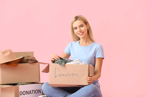 Mujer joven y cajas con ropa para donación sobre fondo rosa - Foto, imagen