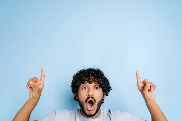 Shocked surprised indian or arabian young man in basic t-shirt, amazed looks at camera with mouth open and points fingers up, at empty space, stands on isolated blue background. Copy-space concept - 写真・画像