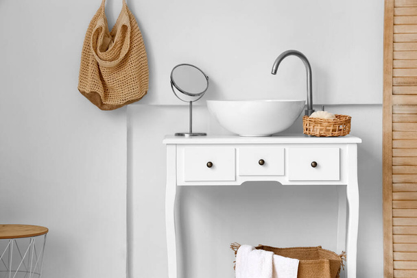 Стол с раковиной, зеркалом и корзиной возле светлой стены в ванной комнате - Фото, изображение