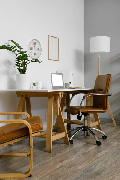 Робоче місце з сучасним ноутбуком і кімнатною рослиною біля світлої стіни в інтер'єрі офісу
 - Фото, зображення