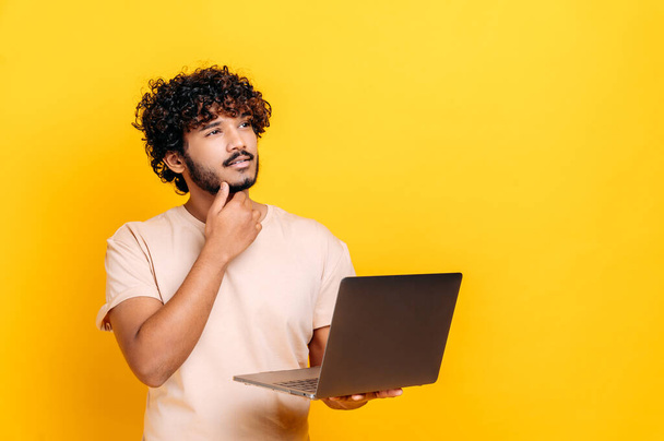 Handsome charismatic indian or arabian curly guy in basic t-shirt, holding open laptop in hand, looking away thoughtfully, planning, dreaming, standing over isolated orange background - Fotografie, Obrázek