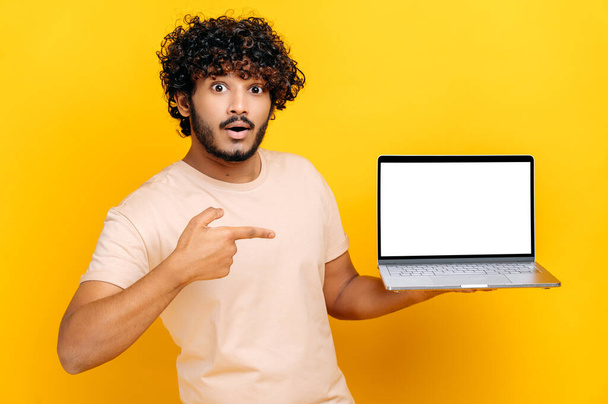 Surprised Indian or Arabian curly guy, holds an open laptop with blank white screen, points finger at it, standing on isolated orange background, confused face expression. Copy space, mock-up concept - Foto, Bild