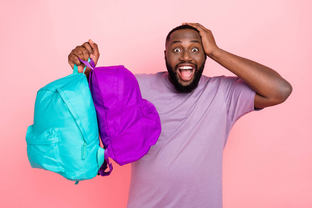 Photo of funny impressed guy wear purple t-shirt rising two schoolbags arm head isolated pink color background - Foto, immagini
