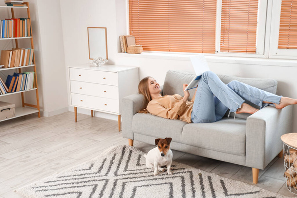 Mujer joven con Jack Russel terrier usando el ordenador portátil en el sofá gris en casa - Foto, imagen