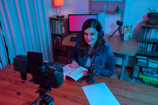 Mooie Spaanse vrouw voor een videocamera die een blog opneemt in haar studio met rood en blauw licht in haar huis. Uitzending van uw commentaar op een boek op sociale media - Foto, afbeelding