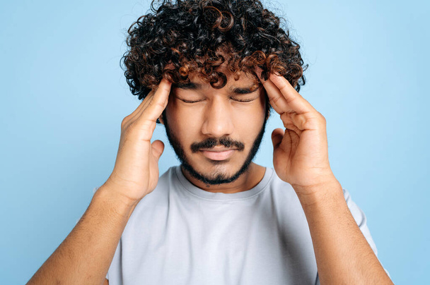 Close-up of sad unhealthy indian guy with eyes closed, massaging his temples, experiencing a migraine, suffering from headache, overworked, in need of rest or treatment, stand isolated blue background - 写真・画像