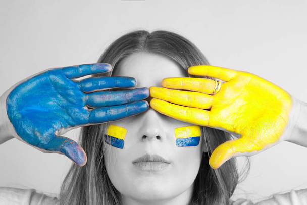 Jovem mulher cobre os olhos com as mãos pintadas na cor da bandeira ucraniana isolada em um fundo cinza. Fotografia em preto e branco. - Foto, Imagem