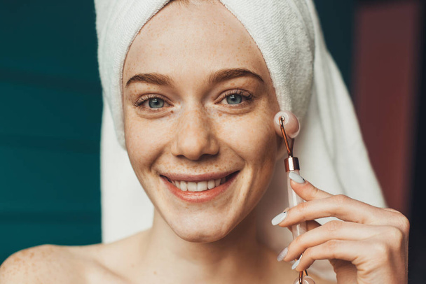 Τζίντζερ γυναίκα με πετσέτα στο πρόσωπο μασάζ κεφάλι χρησιμοποιώντας νεφρίτη ρολό. Ιατρική περίθαλψη. - Φωτογραφία, εικόνα