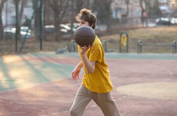 Aranyos fiatal tinédzser sárga pólóban egy labdával kosarazik. Sport, hobbi, aktív életmód fiúknak - Fotó, kép