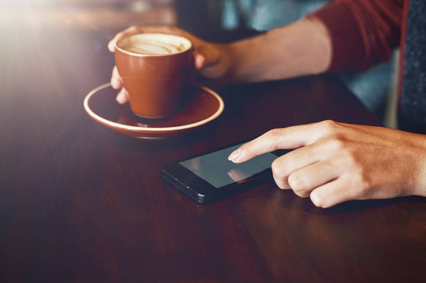 Ik heb gezelschap nodig bij deze koffie. Close-up shot van een niet-identificeerbare vrouw genieten van een kopje koffie tijdens het gebruik van haar telefoon in een cafe. - Foto, afbeelding