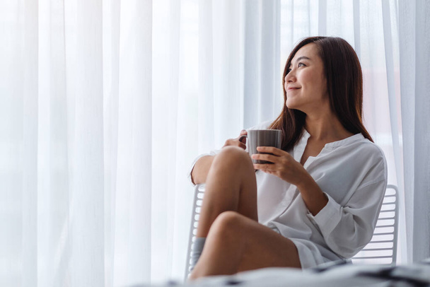 Красива азіатська жінка п'є гарячу каву в білій спальні вдома вранці
 - Фото, зображення