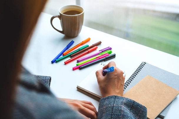 Imagen de cerca de una mujer escribiendo en un cuaderno en blanco con bolígrafos de colores y taza de café en la mesa  - Foto, imagen