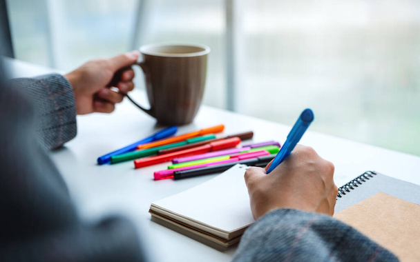 Immagine ravvicinata di una donna che scrive su un quaderno bianco con penne colorate mentre beve caffè  - Foto, immagini