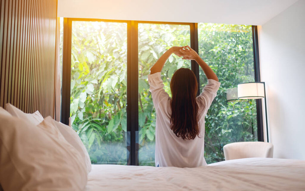 Οπίσθια εικόνα μιας γυναίκας κάνει τέντωμα μετά το ξύπνημα το πρωί, κοιτάζοντας μια όμορφη θέα στη φύση έξω από το παράθυρο του υπνοδωματίου  - Φωτογραφία, εικόνα