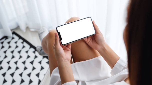 Mockup-Bild der Frau hält Handy mit leerem Desktop-Bildschirm, während sie im Schlafzimmer sitzt und sich entspannt fühlt - Foto, Bild