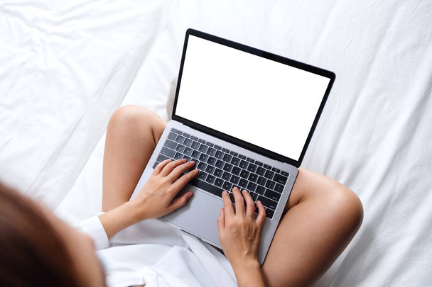 Horní pohled mockup obraz ženy pracující a psaní na notebooku s prázdnou obrazovkou, zatímco sedí na bílé útulné posteli doma - Fotografie, Obrázek
