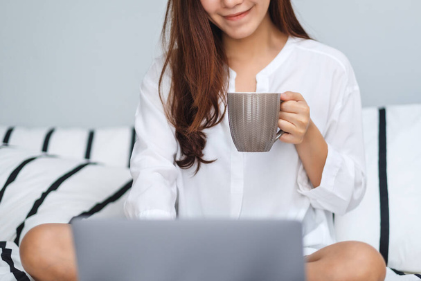 Image rapprochée d'une belle femme utilisant et travaillant sur un ordinateur portable, buvant du café assis sur un lit confortable blanc à la maison - Photo, image
