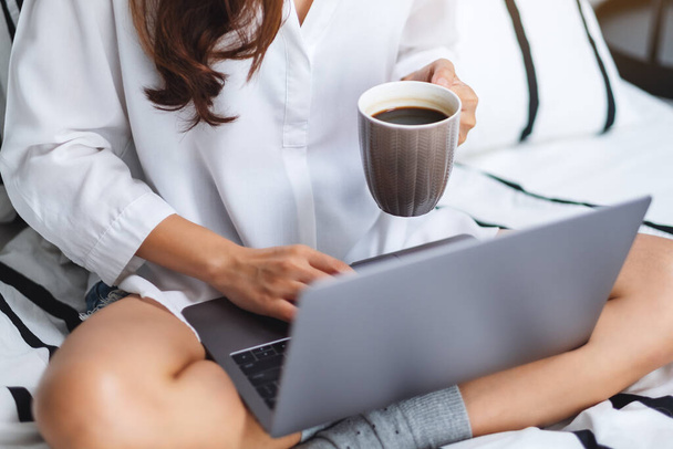 自宅の白い居心地の良いベッドに座っている間にコーヒーを飲みながら、ラップトップコンピュータを使用して作業している女性のイメージを閉じます - 写真・画像