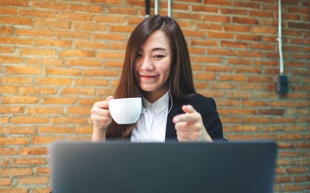 Closeup image of a beautiful businesswoman using earphone for video conference on laptop computer while drinking coffee and working online - Photo, Image