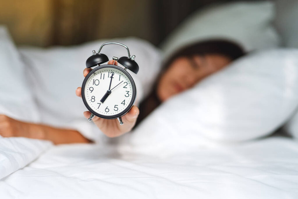 Μια Ασιάτισσα κρατώντας και δείχνοντας ένα ξυπνητήρι ενώ κοιμάται σε ένα λευκό άνετο κρεβάτι το πρωί - Φωτογραφία, εικόνα