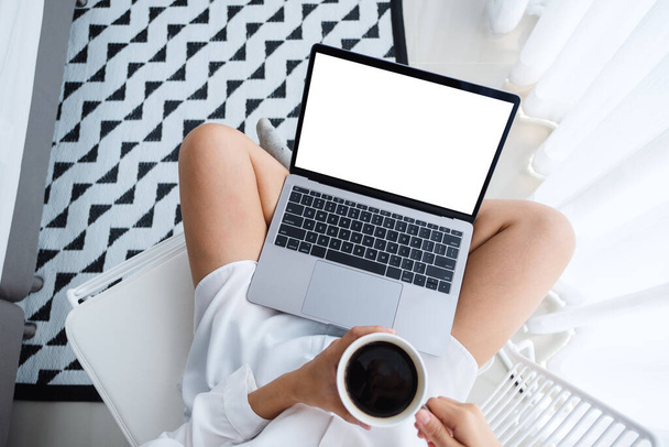Top view mockup afbeelding van een vrouw met behulp van laptop computer met leeg scherm tijdens het zitten en het drinken van koffie in een slaapkamer thuis - Foto, afbeelding