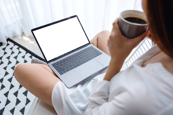 Εικόνα mockup μιας γυναίκας που εργάζεται και δακτυλογραφεί σε φορητό υπολογιστή με κενή οθόνη ενώ κάθεται και πίνει καφέ σε ένα υπνοδωμάτιο στο σπίτι - Φωτογραφία, εικόνα