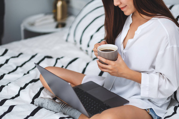 Zbliżenie obrazu kobiety używającej laptopa i pracującej na nim, pijącej kawę siedząc na białym, przytulnym łóżku w domu - Zdjęcie, obraz