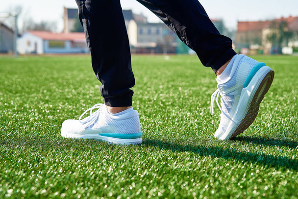 Férfi futólábak fehér tornacipővel a stadionban zöld fűvel, közelről. Sportcipő kocogáshoz. Fitness és egészséges életmód koncepció - Fotó, kép