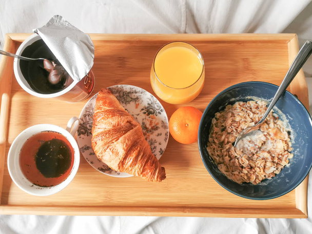 Snídaně v posteli na skládacím dřevěném stole s croissantem, pomerančovým džusem, šálkem čekanky, chokolátovým pomazánkou a müsli. - Fotografie, Obrázek