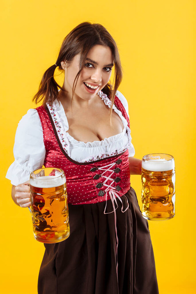 Beautiful waitress wearing traditional dirndl and holding two mass beer steins over yellow background. - 写真・画像