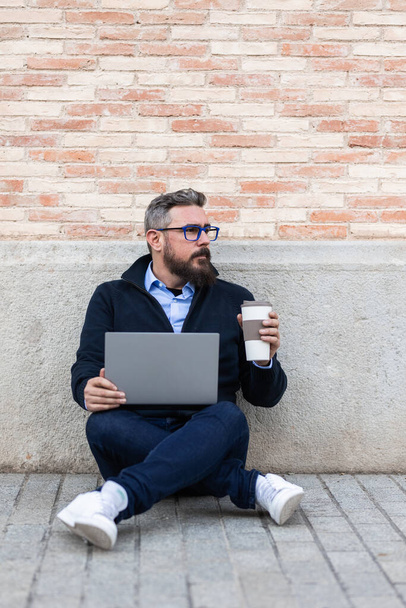 Επιχειρηματίας που εργάζεται με ένα φορητό υπολογιστή και πίνοντας καφέ, ενώ κάθεται σε εξωτερικούς χώρους στο δρόμο. - Φωτογραφία, εικόνα