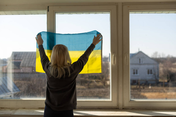 Ukrayna bayrağına sarılmış bir kadın pencereden dışarı bakıyor. Boşluğu kopyala - Fotoğraf, Görsel