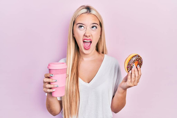 Young blonde girl eating doughnut and drinking coffee angry and mad screaming frustrated and furious, shouting with anger looking up.  - Foto, immagini