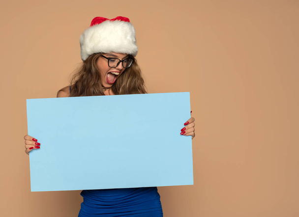 Χριστούγεννα, Χριστούγεννα, άνθρωποι, διαφήμιση, πώληση έννοια - ευτυχισμένη γυναίκα σε μπλε φόρεμα και Σάντα καπέλο βοηθός με κενό μπλε πίνακα σε ένα μπεζ φόντο - Φωτογραφία, εικόνα