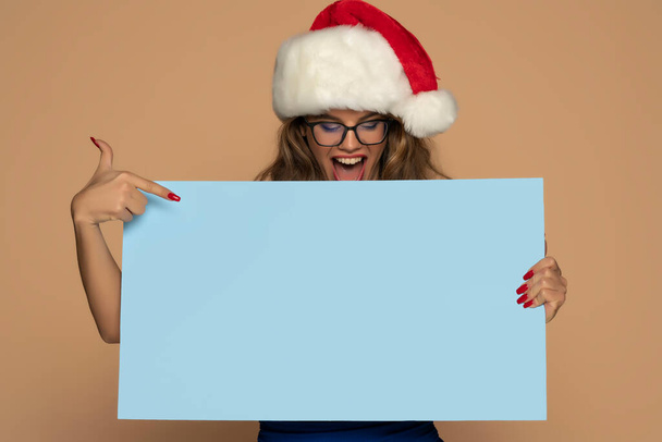 Noel, Noel, Noel, insanlar, reklam, satış konsepti - mavi elbiseli mutlu kadın ve bej arka planda boş mavi panelli Noel Baba yardımcısı şapkası - Fotoğraf, Görsel