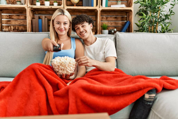 Młoda para ogląda film i je popcorn siedząc na kanapie w domu. - Zdjęcie, obraz